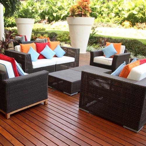 Outdoor seating area with wooden padded chairs with colored pillows on top sitting on lighter wooden flooring around large potted flowers and plants