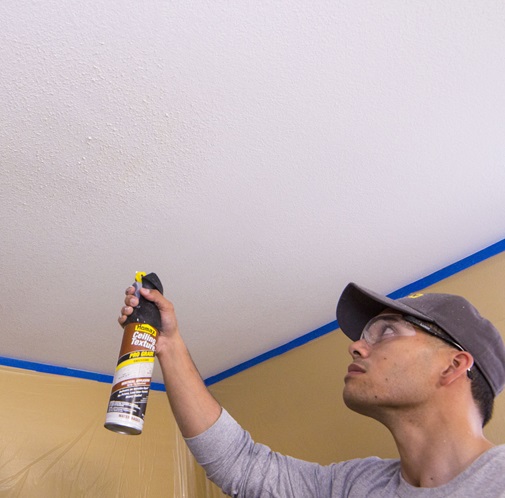 Man with gray hat, shirt, and clear protective glasses looking up at white ceiling holding up spray