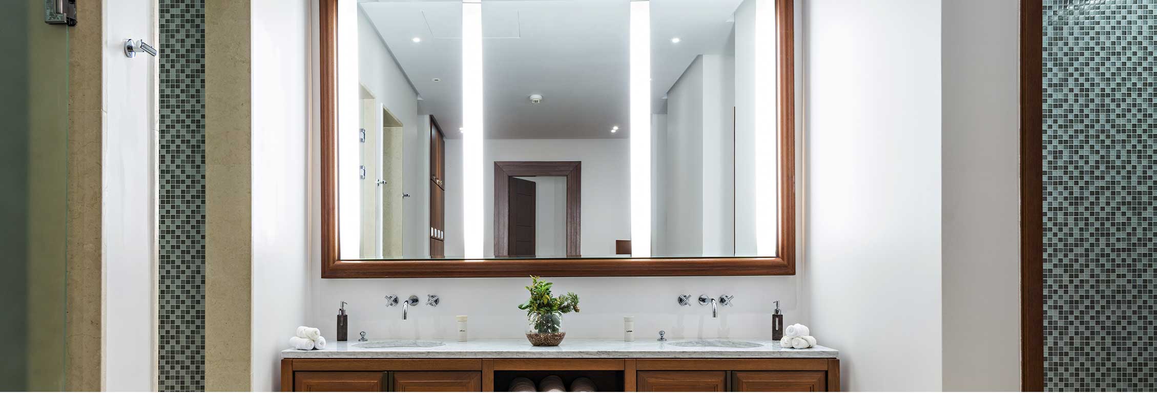 Walls and counter with double sink and large mirror reflecting rest of the white-walled and brown-framed room