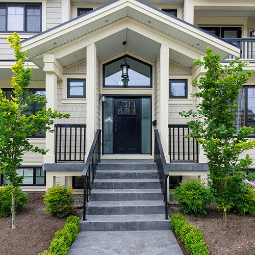 Cream color house with black framing, railings, and door with gray steps and walkway with bushes and tall plants