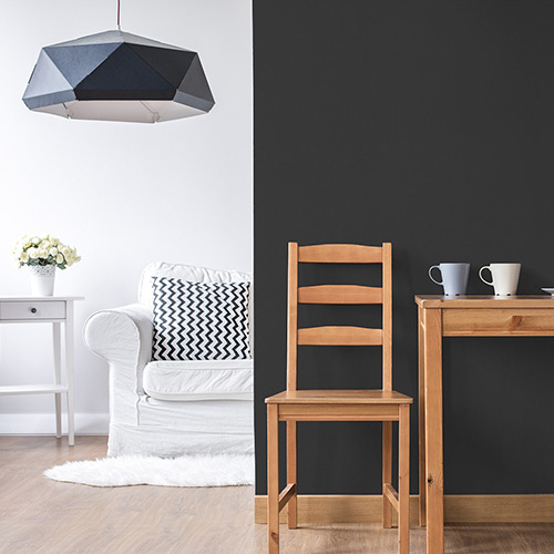 Room with black wall with brown wooden chair and table on right, and white wall with white couch, gray pillow, black decorative hanging light, white side table and flowers on left in background