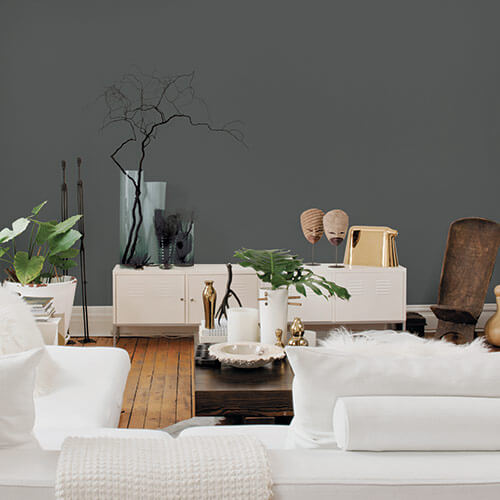 Livingroom with white couches, gray wall, and white side tables with brown wooden flooring and coffee table, with potted plants and wooden decoratives in background and around