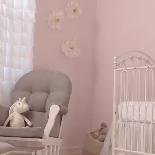 Bedroom with light pink walls and decorative flowers with white crib on right and white curtain, gray chair, and stuffed unicorn on left
