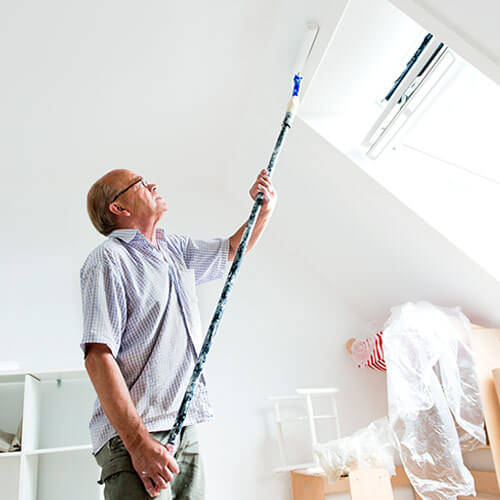 Older man using extended paint roller to paint above slanted window in white room