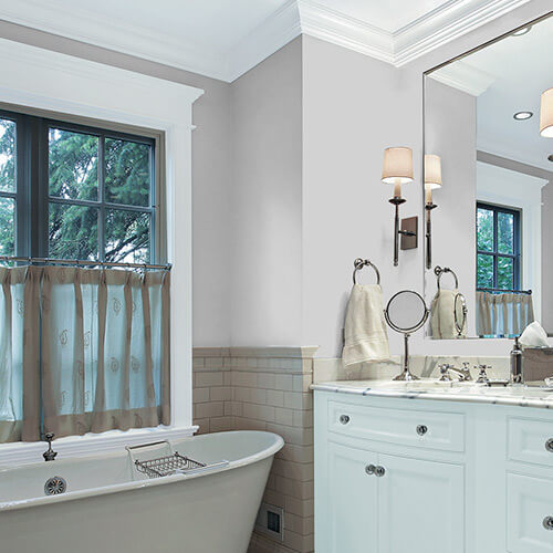 Bathroom with light gray wall on top and tan brick on bottom with white sink and bathtub, mirror, large half-curtained window and small accessories on sink-top