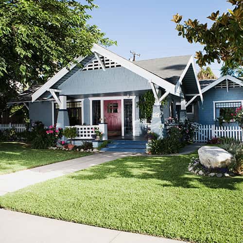 House with light blue panel walls, gray roof, white framing and red door with a front lawn, walkway, and trees around