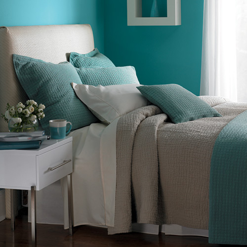 Bedroom with cyan walls, bed with white, gray, and cyan colors, white side table with plant, mug, and small items with painting on cyan walls and window on right