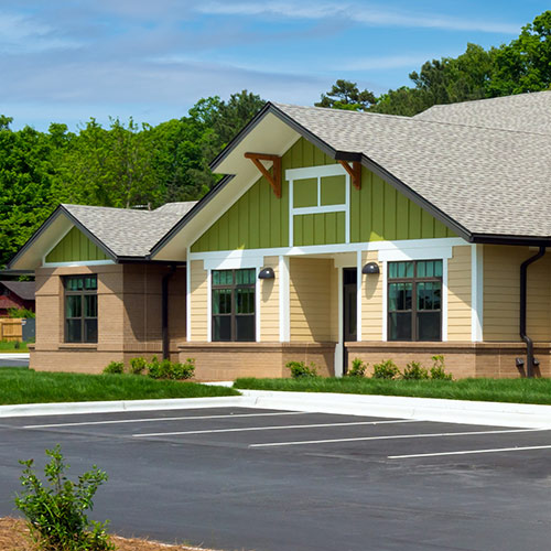 Pale house with gray roof and white framing, light brown bottom brick, parking lot at bottom and trees in background