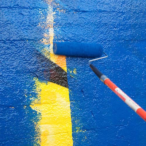 Extended paint roller painting blue paint over yellow and black wall