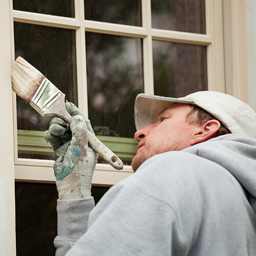Man wearing hat, hoodie, and gloves painting next to framed window