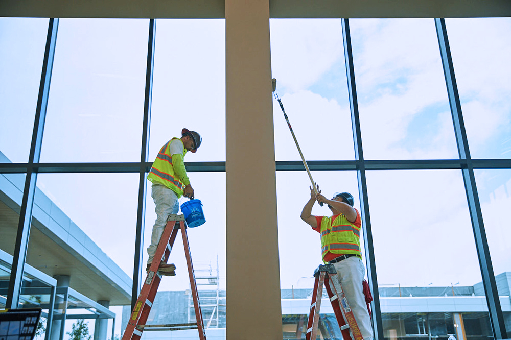 Two men in bright construction gear on ladders using extended rollers to paint tall pillar next to giant windows 