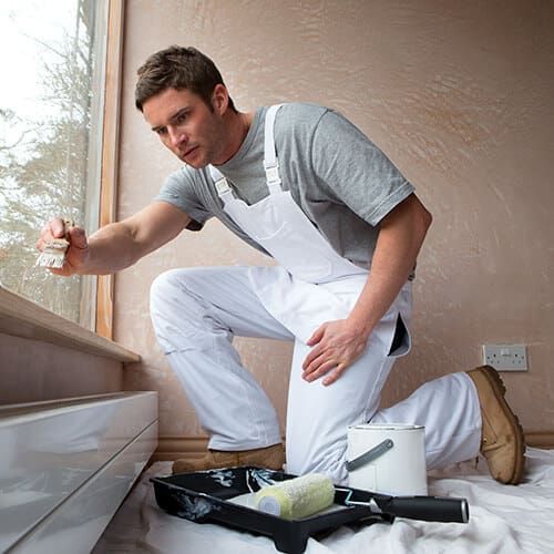 Man with gray shirt and white overalls kneeling and painting with roller and white paint bucket on ground with protective floor covering and tan wall with large window on left