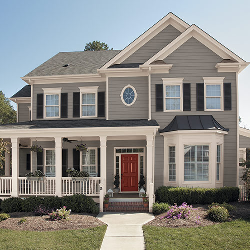 Gray house with gray roof, white framing, black shutters on many windows with bright red door, front lawn with bushes and walkway