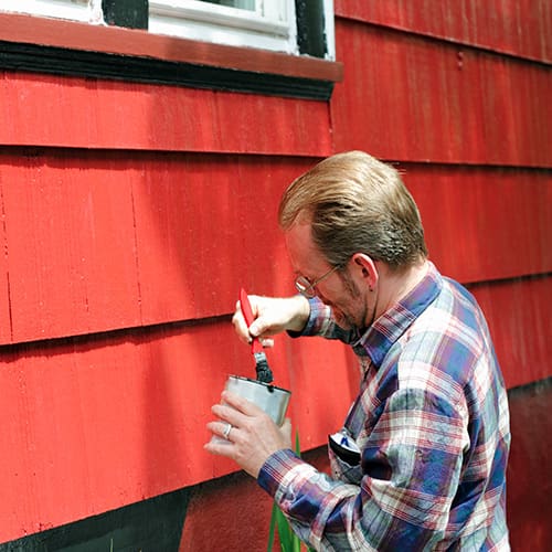 Man dipping paint brush to paint red on house panels