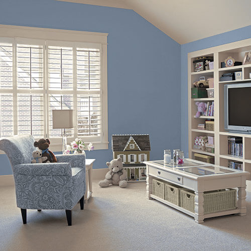 Children's room with blue walls and white carpet and furniture with blue designed chair, large windows, brown mini house and other stuffed animals and toys