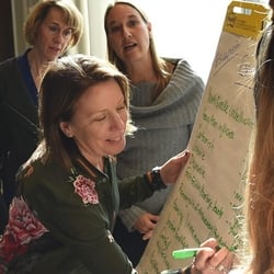 Woman writing on whiteboard with green marker with two women looking behind her 