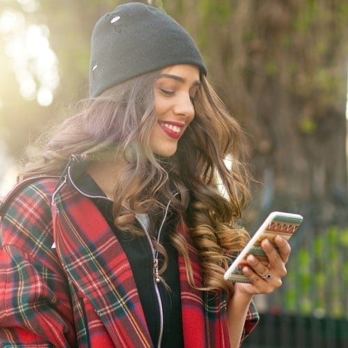 Woman with long brown hair with black beanie and red flannel smiling and holding phone with blurred forest in background