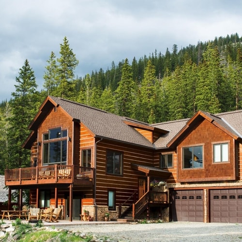 Brown wooden house with roof with windows and brown garages with stone driveway and forest in background