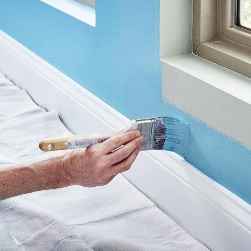 Person's hand holding paint brush painting light blue on wall above white framing and protective floor cover