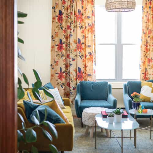 Livingroom with white wall, blue and yellow couch, chairs, and pillows, glass coffee table and large window with flower curtains