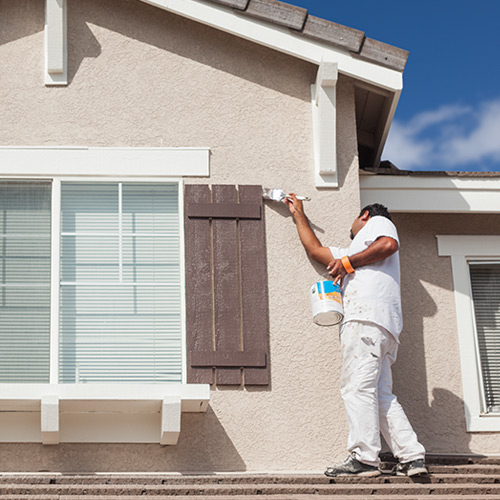 Man wearing all white standing on first story roof painting window shutters dark brown