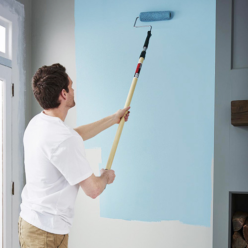 Man with dark hair and white shirt painting light blue onto elongated paint roller on white wall