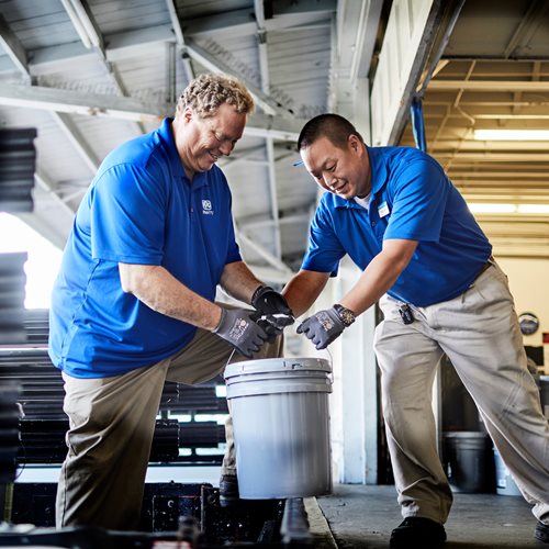 One man with dirty blonde hair and one with short black hair both wearing blue and tan outfits holding the same gray bucket handle in warehouse area
