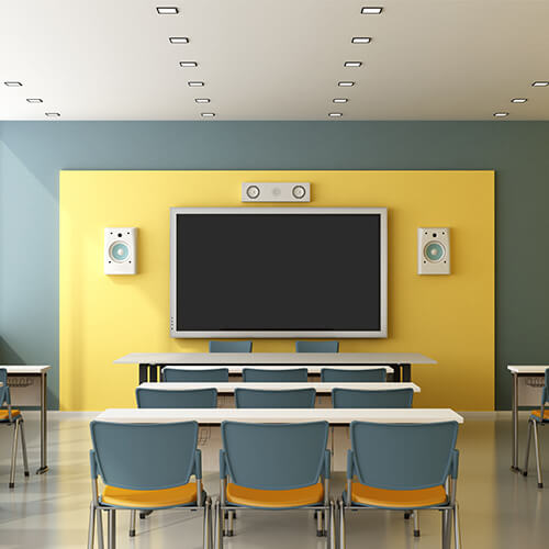 Conference-like room with gray and yellow wall, white ceiling and gray floor, long white desks with gray and orange chairs with large TV on wall with white speakers around