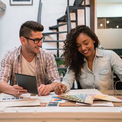 Man with dark hair and glasses holding tablet with woman with dark hair looking at color swatches on table with staircase behind them