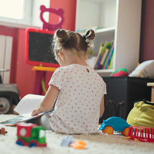 Little girl facing away with buns in hair and white shirt sitting on ground with colorful toys in pink walled room