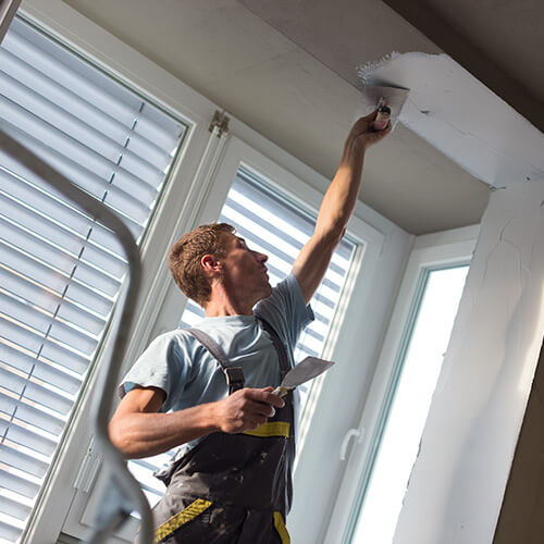 Man with overalls reaching up to paint white paint on ceiling arch next to window