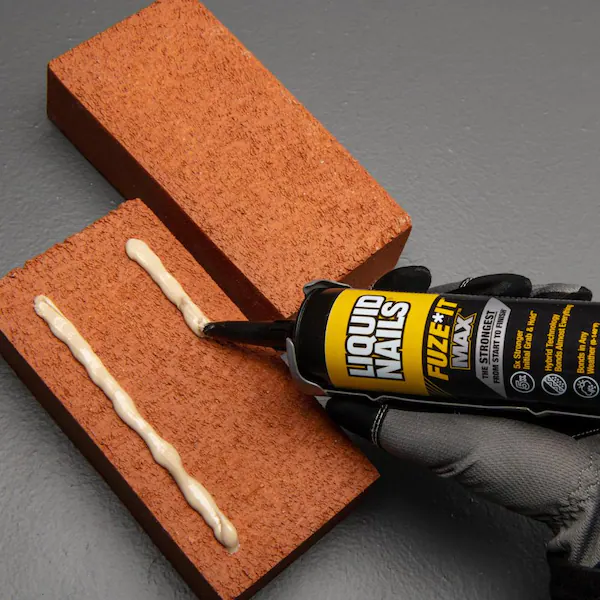 Person wearing gloves holding Liquid Nails adhesive putting liquid on block of wood on gray flooring