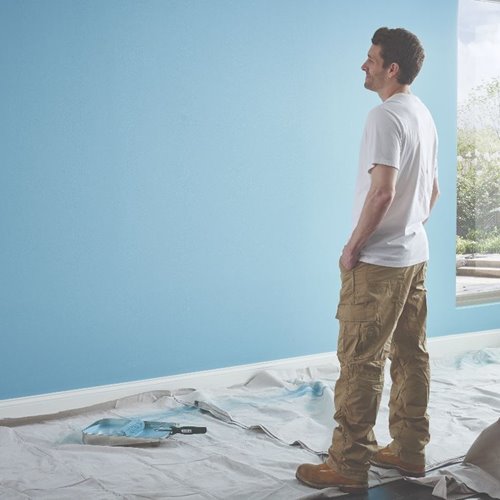 Man with white shirt and tan pants looking at light blue wall standing on protective tarp